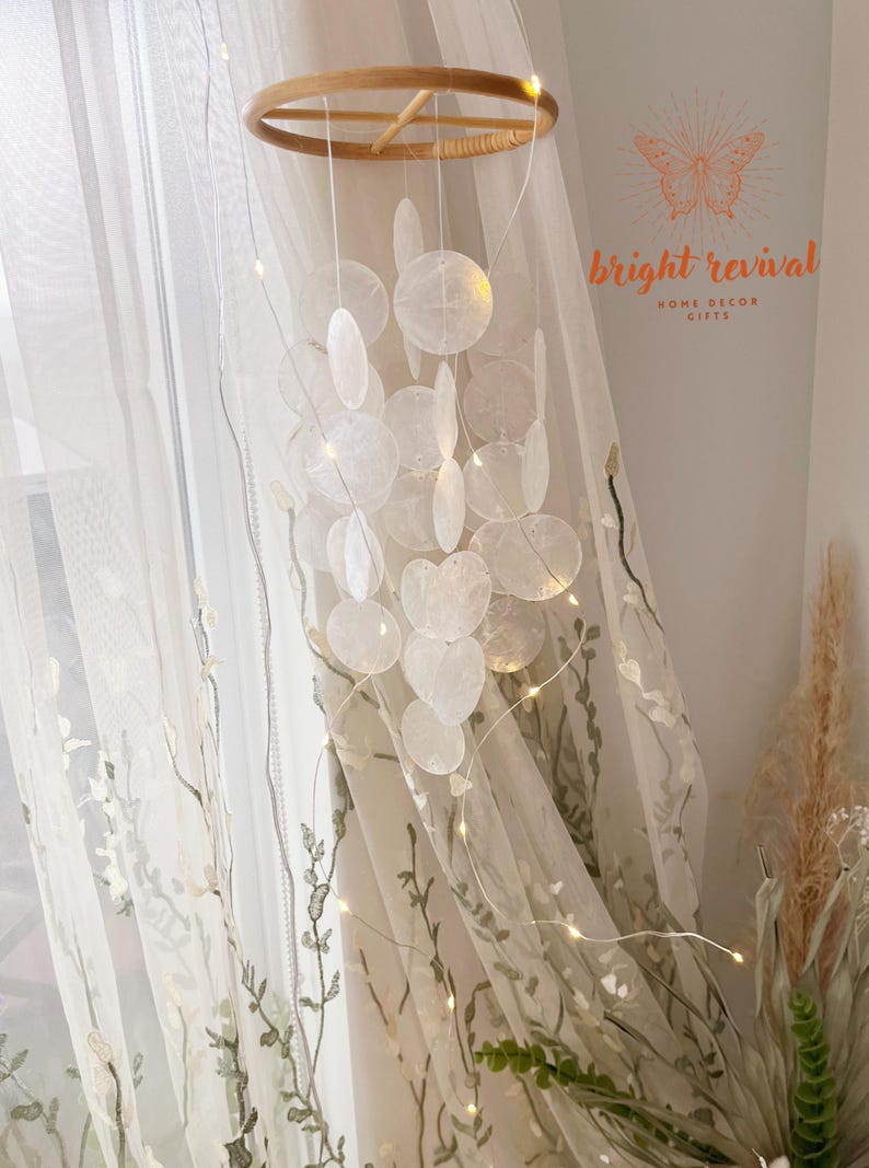 May include: A decorative wind chime featuring iridescent, shell-shaped pendants suspended from a wooden ring. The chime is illuminated by small, warm white fairy lights. The background includes sheer curtains and the text "bright revival HOME DECOR GIFTS".