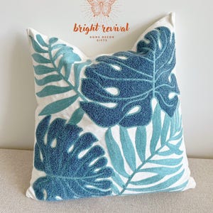 Puede incluir: Un cojín cuadrado blanco con hojas tropicales bordadas en azul y turquesa. El cojín presenta un gran diseño de hoja de monstera texturizada y otros patrones de hojas. Es un artículo de decoración del hogar.