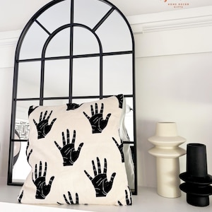 May include: A black and white throw pillow with a hand print pattern. The pillow is sitting on a white shelf in front of a black arched window mirror. There are two decorative vases on the shelf, one white and one black.