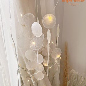 May include: A decorative hanging mobile featuring a natural wood ring and strings of white, translucent, circular and oval-shaped elements. Tiny fairy lights are strung throughout the mobile, illuminating the design. The text "bright revival" is visible.