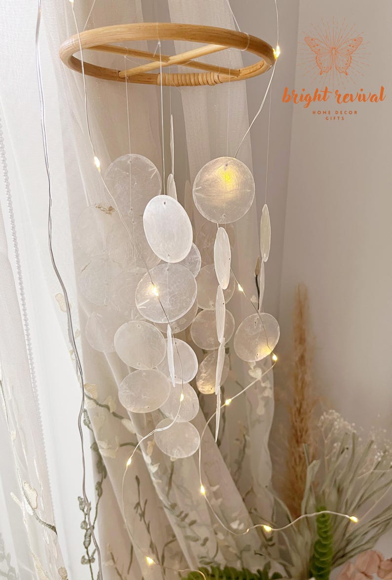 May include: A decorative hanging mobile featuring a natural wood ring and strings of white, translucent, circular and oval-shaped elements. Tiny fairy lights are strung throughout the mobile, illuminating the design. The text "bright revival" is visible.