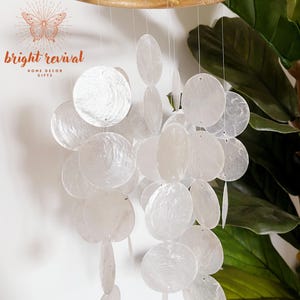 May include: A white capiz shell wind chime with a natural bamboo ring. The chime features multiple round and oval-shaped shells suspended by thin strings. The text "bright revival" is visible in the upper left corner. Home decor.