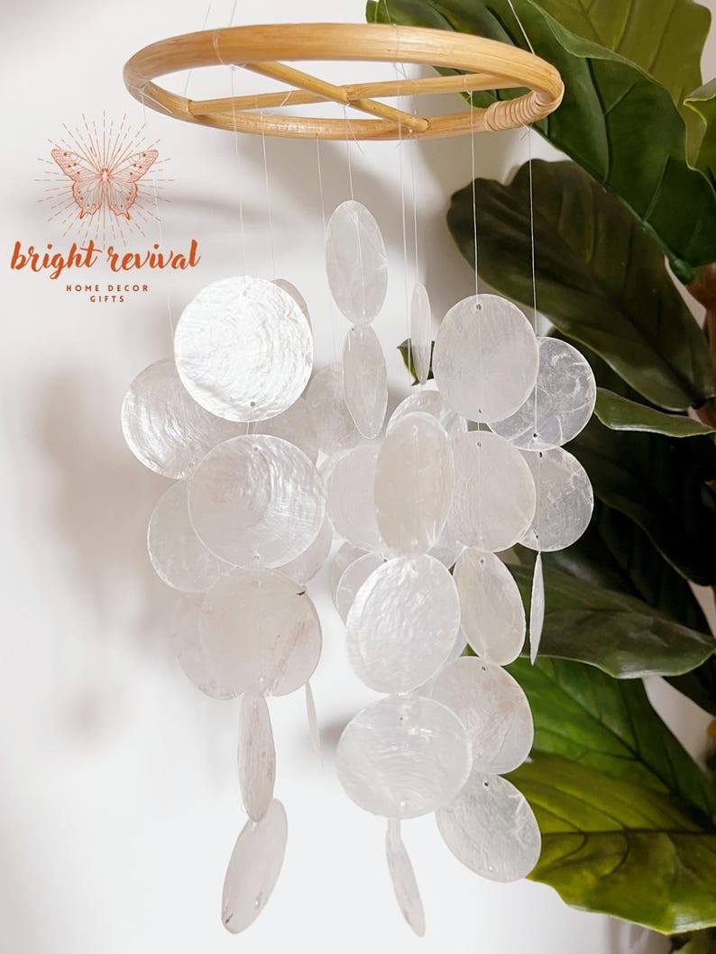 May include: A white capiz shell wind chime with a natural bamboo ring. The chime features multiple round and oval-shaped shells suspended by thin strings. The text "bright revival" is visible in the upper left corner. Home decor.