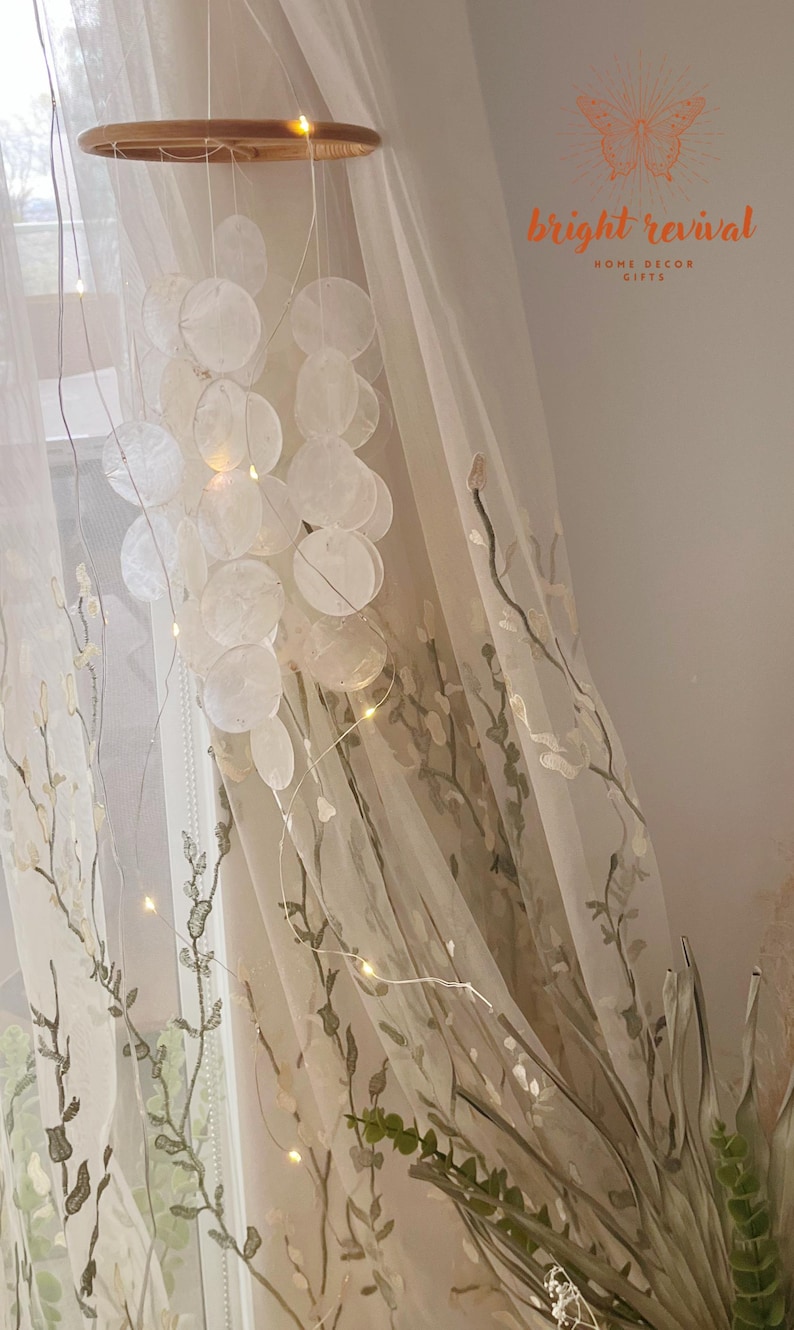 May include: A decorative hanging mobile featuring a wooden ring, strings of small, round, white shell-like discs, and tiny fairy lights. The mobile hangs in front of a sheer curtain with embroidered floral patterns. The text "bright revival HOME DECOR GIFTS" is visible.