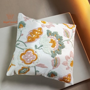 Peut inclure: Coussin décoratif carré blanc avec un motif floral brodé. Le coussin présente un motif de fleurs et de feuilles dans des tons de jaune, rose et vert. Le coussin est posé sur une surface blanche.