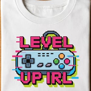 Może przedstawiać: Biała koszulka z pikselowym wzorem kontrolera do gier wideo z różowym i zielonym napisem "LEVEL UP IRL". Kontroler jest jasnoniebieski z kolorowymi przyciskami. Koszulka jest złożona.