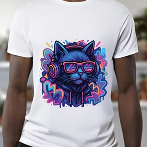 May include: White t-shirt featuring a vibrant graphic of a blue cat wearing headphones and pink sunglasses. The cat is set against a colorful, abstract background with splashes of pink, blue, yellow, and purple. The design has a modern, cartoonish style.