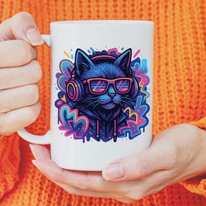 May include: White ceramic mug featuring a black cat wearing headphones and neon pink glasses. The cat is set against a colorful, graffiti-style background. The mug is held by a person wearing an orange sweater.
