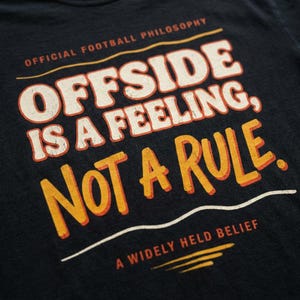 Puede incluir: Camiseta azul marino oscuro con la frase "OFFSIDE IS A FEELING, NOT A RULE" en letras blancas y naranjas. La camiseta también incluye las frases "OFFICIAL FOOTBALL PHILOSOPHY" y "A WIDELY HELD BELIEF".