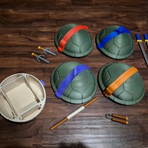 May include: A collection of Teenage Mutant Ninja Turtles cosplay accessories, including four green turtle shells with colored headbands, weapons, and a shell-shaped container. The weapons include swords, sais, and nunchucks.