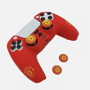 May include: Red and white PlayStation 5 controller with Manchester United logo and thumbstick covers. The controller has a red silicone skin with the Manchester United crest on the back. The thumbstick covers are also red with the Manchester United crest.