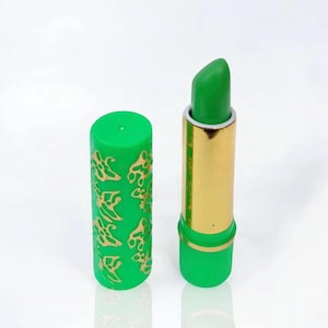 May include: Green lipstick in a gold tube with a green cap. The cap features a gold floral pattern. The lipstick itself is a vibrant green. The lipstick is on a white surface.