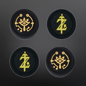 May include: Four black round buttons with gold designs. Two buttons feature a stylised eye with a triangle and a circle around it. The other two buttons feature a stylised letter Z with a cross and a star.