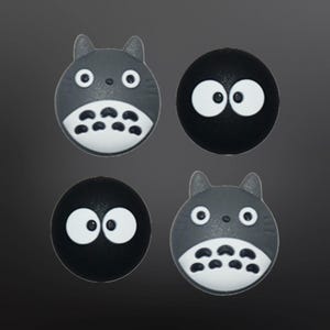 May include: Four round, grey and black, cartoon character-shaped, silicone buttons. Two buttons feature a grey cat-like character with large eyes and a white mouth. The other two buttons are black with white eyes.