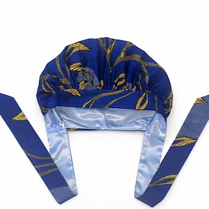 May include: A blue bonnet featuring a yellow leaf pattern on the exterior and a smooth, light blue satin lining. The head covering includes two long, matching fabric ties extending from the sides, designed to secure the cap around the head.