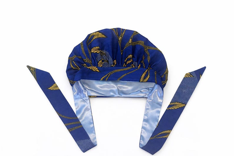 May include: A blue bonnet featuring a yellow leaf pattern on the exterior and a smooth, light blue satin lining. The head covering includes two long, matching fabric ties extending from the sides, designed to secure the cap around the head.