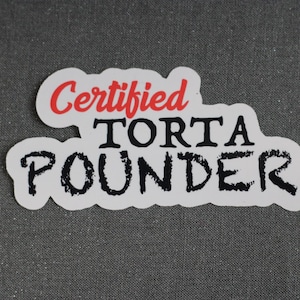 May include: A white sticker with the words "Certified Torta Pounder" in black and red lettering. The sticker has a unique cloud-like shape and is set against a gray background.