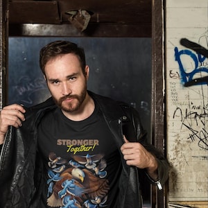 Puede incluir: Una camiseta negra con un gráfico de un águila calva rodeada de pájaros azules. El texto "Stronger Together!" está impreso en amarillo en la camiseta.