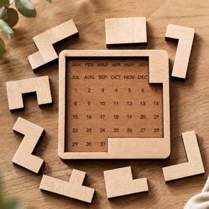 Op de afbeelding: Een houten eeuwigdurende kalender met een vierkant frame en een kalenderraster. De kalender is omgeven door verschillende houten puzzelstukjes. De maanden en data staan op de kalender gedrukt. De kalender ligt op een houten oppervlak.