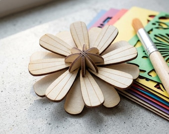 May include: A light-colored wooden flower craft piece with layered petals, featuring a central interlocking structure. Beside the flower are several colorful cardstock sheets with intricate cut-out patterns and a wooden-handled paintbrush.