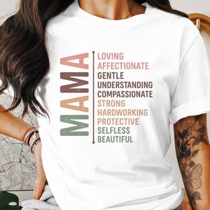 Op de afbeelding: Wit T-shirt met het woord "MAMA" in groene, bruine en roze letters. De letters staan verticaal op elkaar en elke letter heeft een woord ernaast: Loving, Affectionate, Gentle, Understanding, Compassionate, Strong, Hardworking, Protective, Selfless, Beautiful.