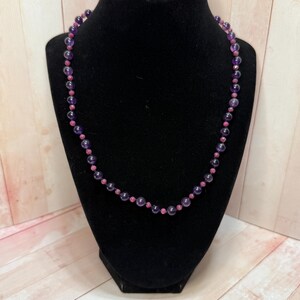 Pode incluir: Um colar de contas roxas e cor-de-rosa em um expositor preto. O colar é feito de contas redondas, sendo as contas roxas maiores que as cor-de-rosa.