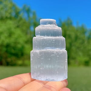 May include: A light blue selenite tower, a crystal formation with a layered design. The tower is held in a hand, with a blurred green and blue background.