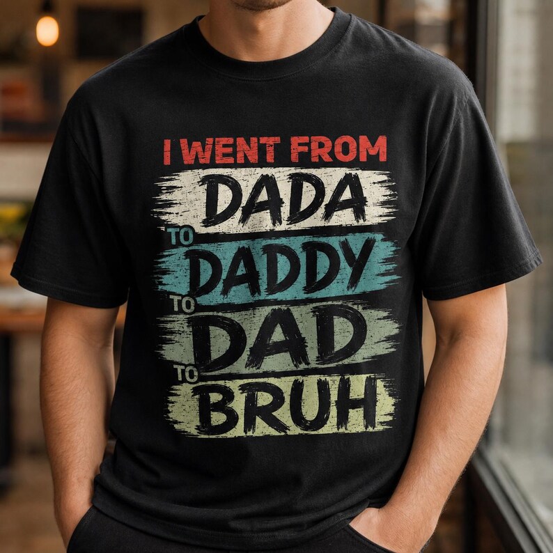 May include: Black t-shirt with the text "I WENT FROM DADA TO DADDY TO DAD TO BRUH" in a distressed, layered design. The text is in red, white, blue, and green colors. The shirt is a casual wear item.