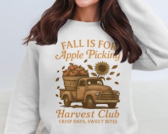 May include: White sweatshirt with a fall-themed graphic. The design features a vintage truck carrying a basket of apples, a sunflower, and the text "FALL IS FOR Apple Picking" and "Harvest Club". Crisp days, sweet bites.