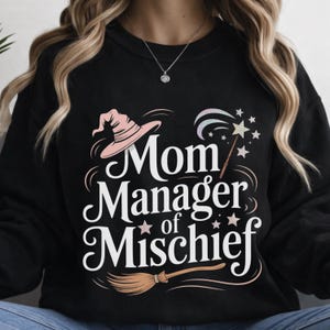 May include: Black sweatshirt with the words "Mom Manager of Mischief" in white script. The design includes a pink witch hat, a magic wand with stars, and a broom. A fun and whimsical design.