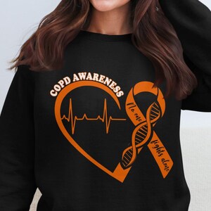 Puede incluir: Sudadera negra con un diseño naranja en forma de corazón que incluye una línea de latidos y las palabras "Concienciación sobre la EPOC". También presenta una cinta naranja con una hebra de ADN y las palabras "Nadie lucha solo".