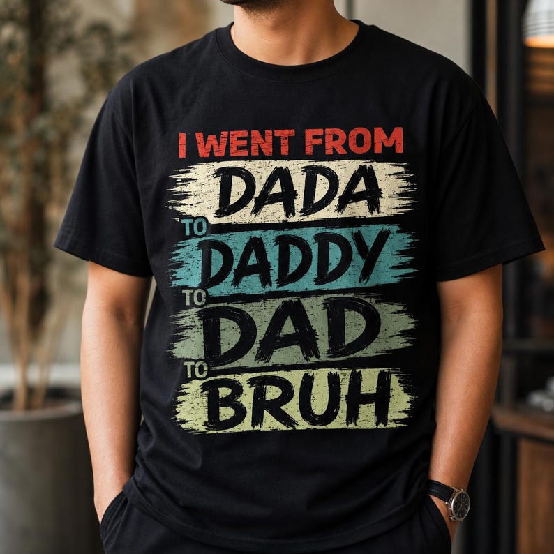 May include: Black t-shirt with the text "I went from Dada to Daddy to Dad to Bruh" in a distressed, brushstroke style. The text is in red, white, blue, and green. The shirt is a casual wear item.