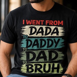 May include: Black t-shirt with the text "I went from Dada to Daddy to Dad to Bruh" in red, white, teal, and green. The text has a distressed, brushstroke style. The shirt is a casual wear item.