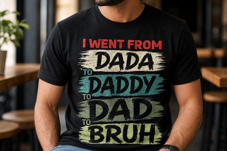 May include: Black t-shirt with the text "I went from Dada to Daddy to Dad to Bruh" in red, white, teal, and green. The text has a distressed, brushstroke style. The shirt is a casual wear item.