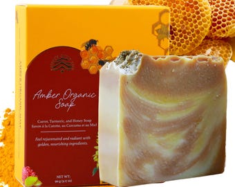 May include: A bar of Amber Organic soap with a swirl of yellow and orange. The soap is packaged in a yellow box with a honeycomb design and the text "Amber Organic Soap" and "Carrot, Turmeric, and Honey Soap".