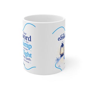 Puede incluir: Una taza de cerámica blanca con texto azul que dice Your word is a lamp to my feet and a light to my path, Psalm 119:105. El diseño incluye un gráfico de una linterna en un camino y texto adicional en una escritura no latina.
