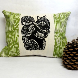Puede incluir: Una almohada verde y blanca con una silueta negra de una ardilla sosteniendo una nuez. La almohada tiene un patrón de veta de madera.