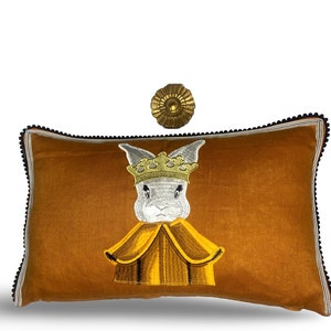 May include: A brown velvet cushion featuring an embroidered white rabbit wearing a gold crown and a yellow robe.