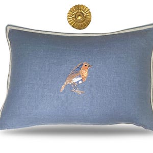 May include: A rectangular blue decorative pillow with a detailed embroidered bird design. The pillow has a white and light blue striped border. A gold-colored sunburst wall decor is above the pillow.