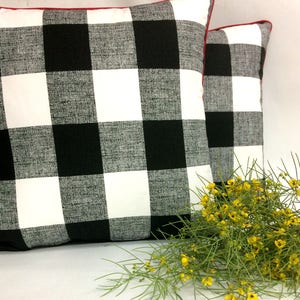 May include: Two black and white buffalo plaid throw pillows with red trim. The pillows are placed on a white surface with a bouquet of yellow flowers in front of them.
