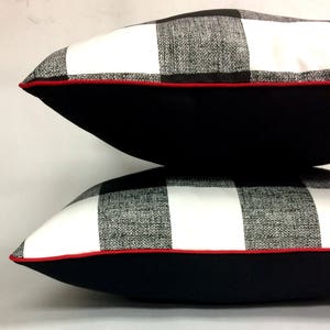 May include: Two black and white checkered throw pillows with red piping. The pillows are stacked on top of each other with the top pillow slightly offset to the right. The pillows have a black backing.