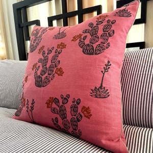 May include: A square decorative pillow with a pink background and a repeating pattern of black and brown cacti. The pillow has a white border and is resting on a striped pillow and a black headboard.