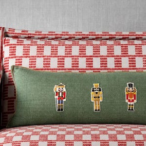 May include: A rectangular olive green lumbar pillow featuring three embroidered nutcracker figures in red, gold, and white uniforms, positioned horizontally across the center of the fabric surface with a piped edge finish.