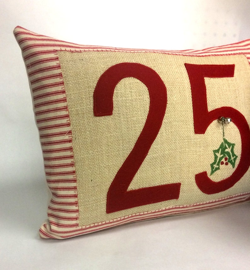 May include: A red and white striped pillow with a burlap front featuring the number 25 in red felt. A small green holly sprig with red berries is attached to the number 5.