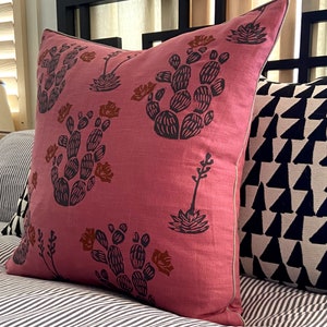 May include: A square, dusty rose pillow with a repeating pattern of dark gray cacti and rust-colored flowers. The pillow has a light gray trim and is placed on a bed with striped bedding. Another pillow with a black and white geometric design is visible.
