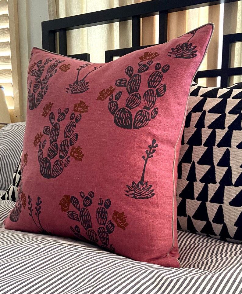 May include: A square, dusty rose pillow with a repeating pattern of dark gray cacti and rust-colored flowers. The pillow has a light gray trim and is placed on a bed with striped bedding. Another pillow with a black and white geometric design is visible.