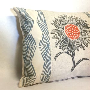 May include: A white pillow with a blue and white geometric pattern on one side and a large orange and gray flower on the other side.