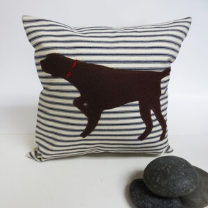May include: A white and blue striped cushion with a brown felt dog silhouette. The dog is wearing a red collar.