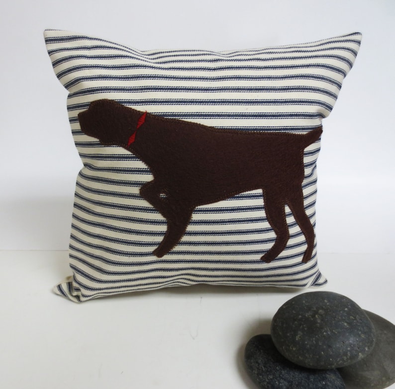 May include: A white and blue striped cushion with a brown felt dog silhouette. The dog is wearing a red collar.