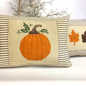 Op de afbeelding: Twee decoratieve kussens met een achtergrond van jute stof. Een kussen heeft een oranje pompoen met groene bladeren en een bruine stengel. Het andere kussen heeft een bruin en oranje blad.